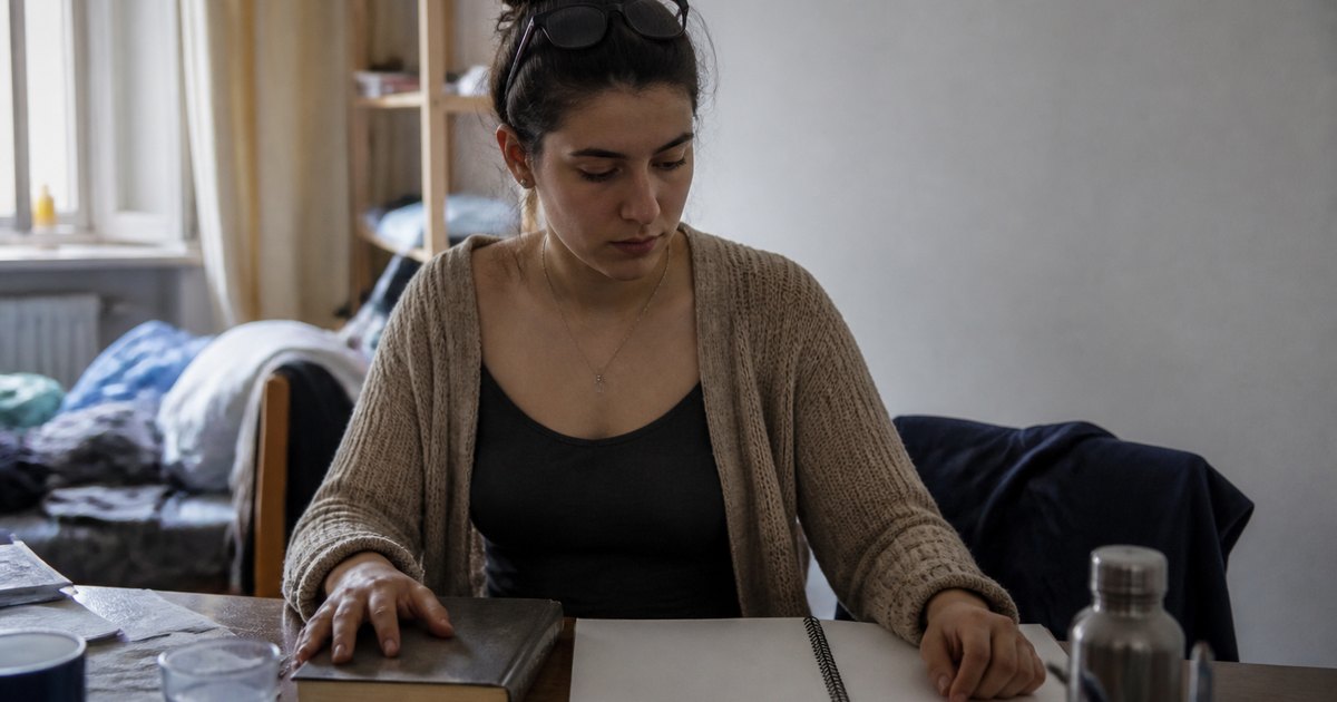 A woman sits at a cluttered desk with a book and blank notebook, looking down in thought.
