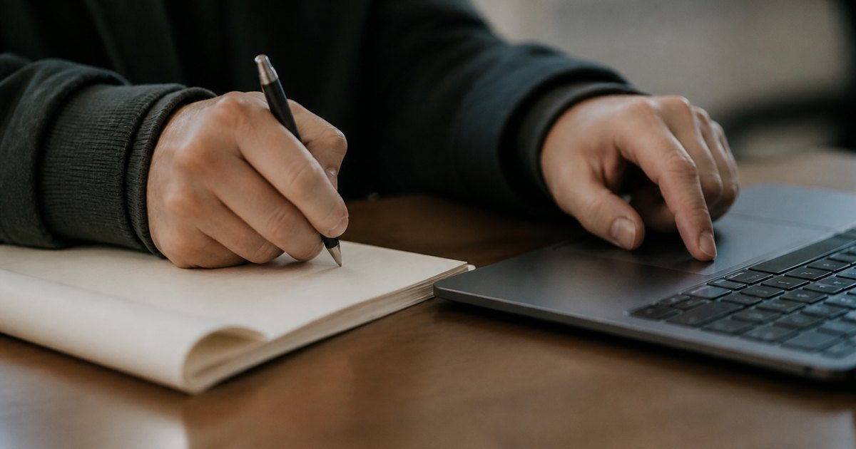 Hands in a coat sleeve use a laptop beside notes and a pen on a study table.