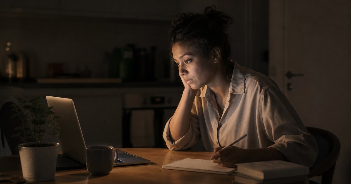 A tired student takes notes from a paused movie on her laptop at a kitchen table.