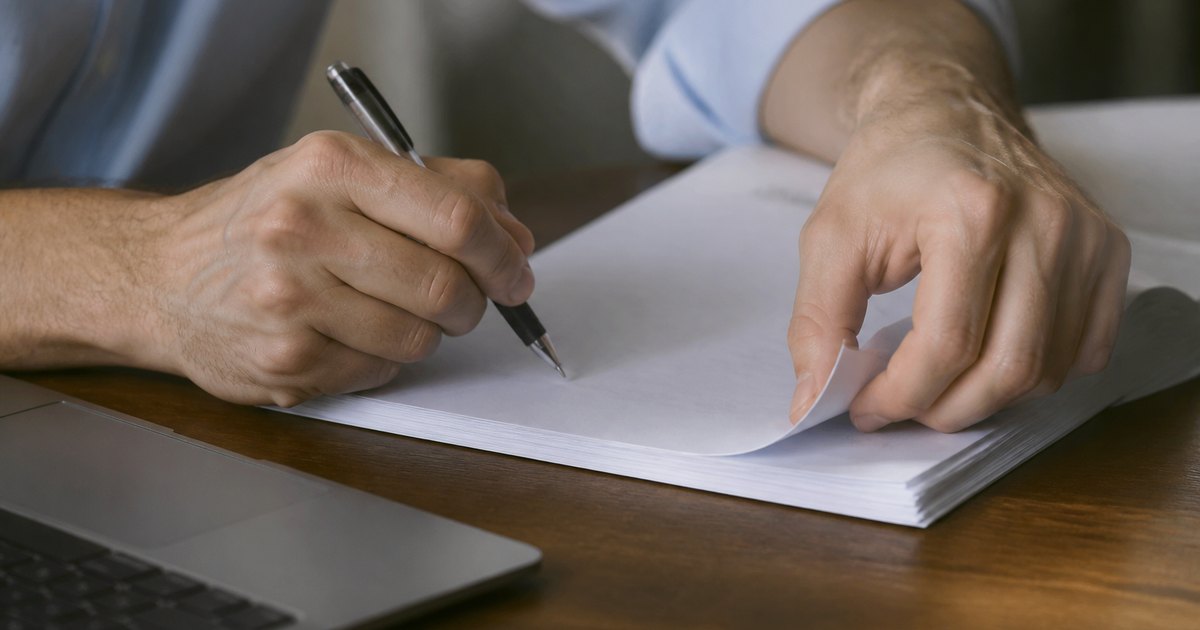Hands proofread a printed page with a pen beside an open laptop.