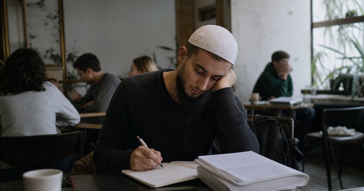 A man writes notes in a notebook at a cafe table with research papers beside him.