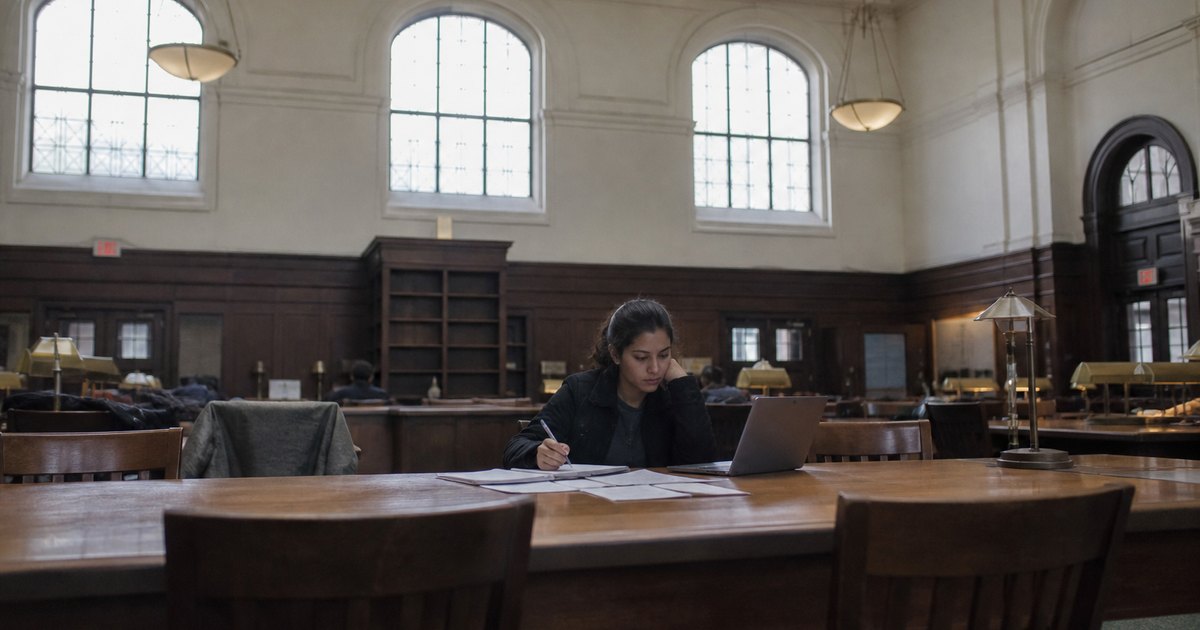A woman sits alone with papers and a laptop in a large library reading room.