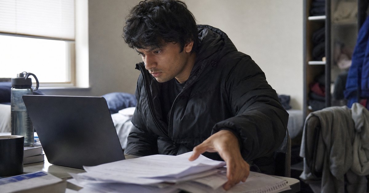 Student at a cluttered desk reviews assignment files on a laptop.