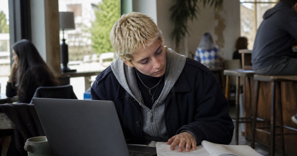 Student at a cafe table reviews an assignment and laptop form while people blur around her.